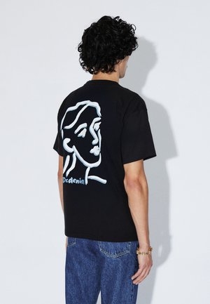 T-shirt en coton noir avec un dessin en ligne blanche représentant un visage au dos. Associé à un jean en denim bleu.