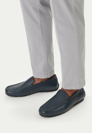 Granatowe skórzane mokasyny wsuwane, noszone z jasnobeżowymi spodniami, pokazane od połowy łydki w dół na białym tle.