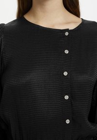 Blusa negra texturizada con escote redondo y seis botones plateados a rayas en el frente. La tela tiene un patrón sutil.