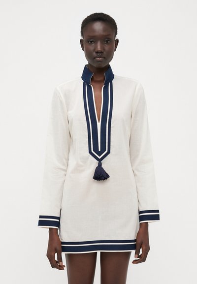 Weißes Tunika-Kleid mit marineblauen Akzenten, ausgestattet mit einem Stehkragen, einem tiefen V-Ausschnitt und einer dekorativen marineblauen Quaste am Saum. Lange Ärmel.