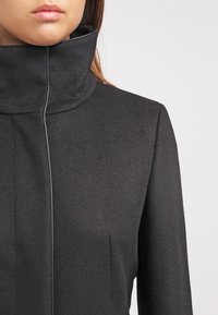 Sort uldblandingsfrakke med høj krave, der har en glat, struktureret overflade og en forhøjet søm. Minimalistisk design uden synlige lukninger.