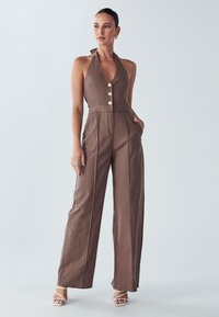 Bruine halter-jumpsuit met een knoopsluiting aan de voorkant, wijde pijpen en zakken. Heeft een gestructureerde stof en wordt gecompleteerd met witte sandalen met bandjes.