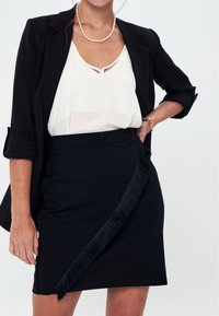 Gonna nera avvolgente con dettagli frangiati lungo il lato, abbinata a una blusa bianca con scollatura a V e un blazer nero. Tessuto morbido, vestibilità su misura.