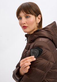 Bruine gewatteerde jas met een capuchon, voorzien van een gestructureerd embleem op de schouder. De stof heeft een gladde afwerking en opgezwollen delen voor isolatie.