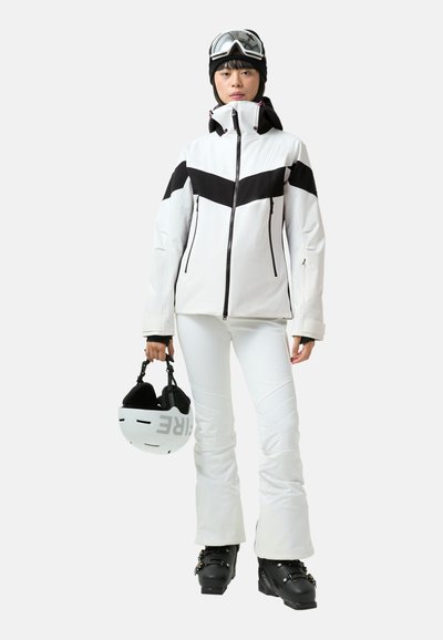 Hvid skijakke med sorte accenter, lynlåslukninger og høj krave, parret med hvide bukser og en hvid hjelm. Modellen holder hjelmen.