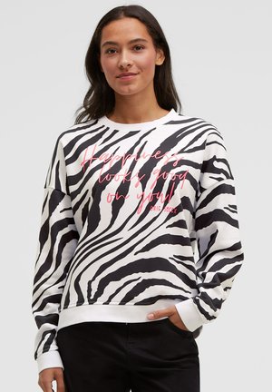 Frau trägt ein schwarz-weißes Sweatshirt mit Zebramuster und pinker Aufschrift "Happiness looks good on you" sowie eine schwarze Hose, Hand in der Tasche.