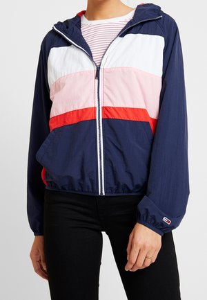Person iført en color-block lynlåstrøje med marineblå, hvid, pink og røde striber og sorte bukser, med en stribet trøje under.