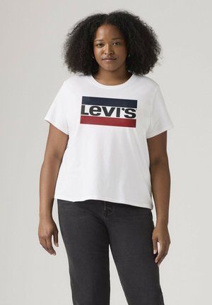 Frau mit lockigem Haar, die ein weißes Levi's T-Shirt mit schwarzen und roten Streifen und schwarze Jeans trägt, steht vor einem schlichten Hintergrund.