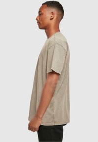 Lys brun, oversized bomulds T-shirt med rund halsudskæring, korte ærmer og rå kantdetaljer, båret af en person i profil.