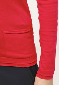 Punainen pitkähihaisen pusero, joka on valmistettu pehmeästä neulekankaasta. Siinä on slim fit -leikkaus, ribbireunukset ja pieni etutasku. Saumadetail hemse kohdalla.