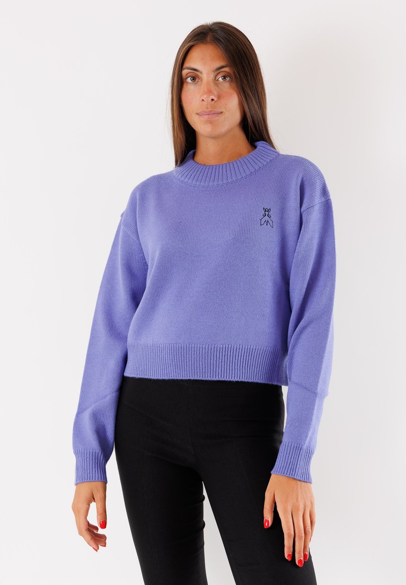 Maglione corto color lilla chiaro con collo e polsini a costine. Presenta un piccolo logo nero sul lato anteriore sinistro. Indossato con pantaloni neri.