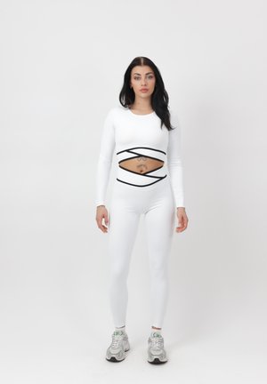 Vrouw staat recht voor zich kijkend, draagt een witte strakke outfit met zwarte kruislings lopende banden die de buik blootlaten, en witte sneakers, tegen een effen achtergrond.