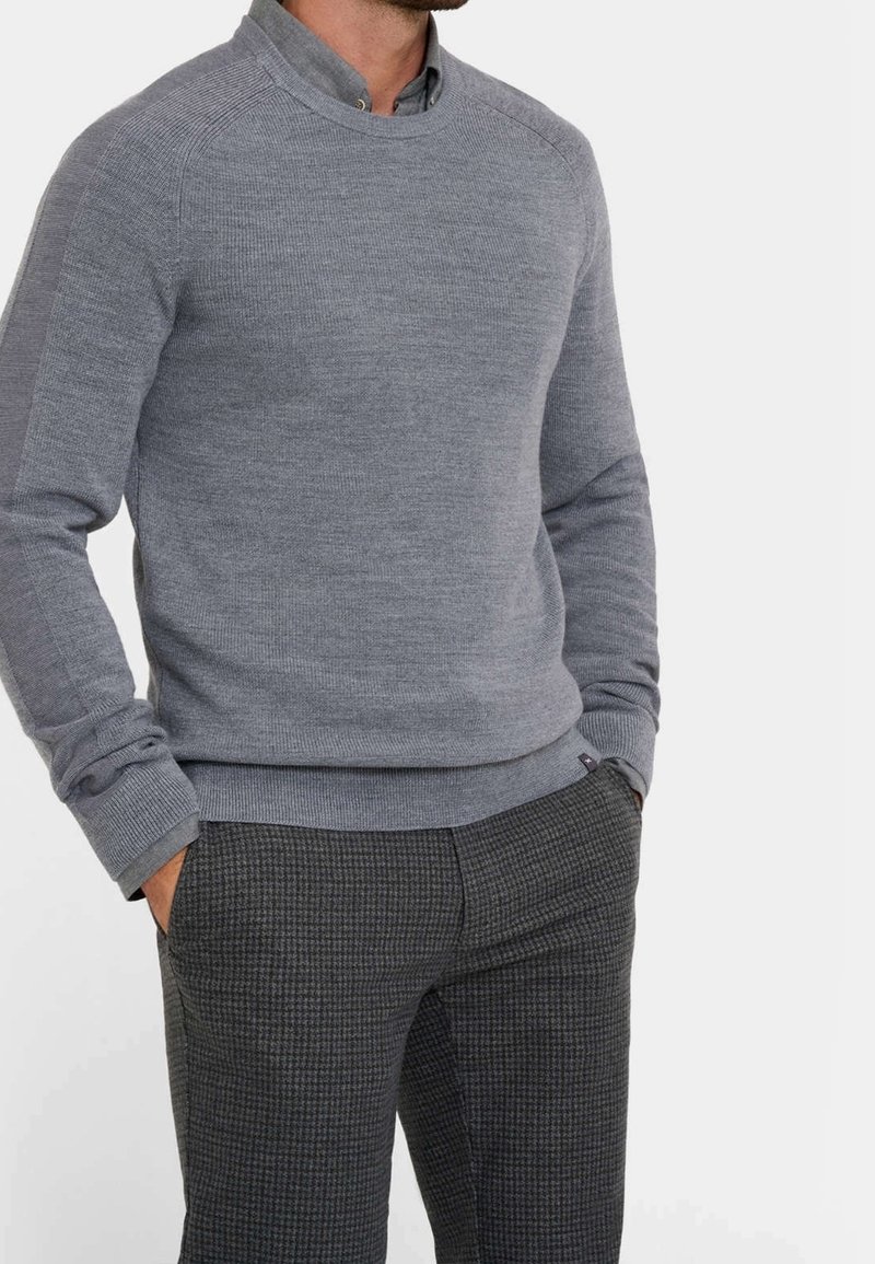 Homme portant un pull gris clair à col rond superposé à une chemise grise à col et un pantalon gris foncé à motif, les mains dans les poches.