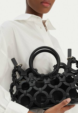 Osoba w białej koszuli trzyma czarną torebkę z okrągłym wzorem wycięć i okrągłymi uchwytami na jednolitym tle.