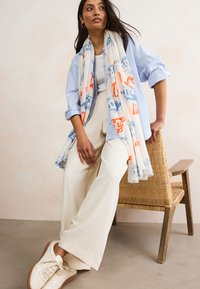 Hellblaues Hemd, cremefarbene Hosen mit weitem Bein und ein gemusterter Schal in Weiß, Blau und Orange. Beige Turnschuhe. Geflochtener Stuhl im Hintergrund.