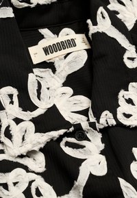 Tissu noir texturé avec des motifs floraux brodés en blanc et une étiquette de la marque Woodbird cousue à l'intérieur du col.