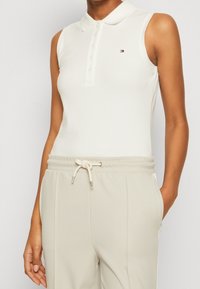 Valkoinen ilman hihoja oleva polo-paita kauluksella ja nappisulkimella, yhdistettynä vaaleanharmaisiin jumppahousuihin, joissa on nauhakuulokkeet.