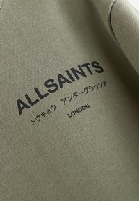 Maslinasto zeleni dukserica s crnim otisnutim natpisom "ALLSAINTS" i "LONDON", uz dodatni tekst na japanskom. Tekstura tkanine.