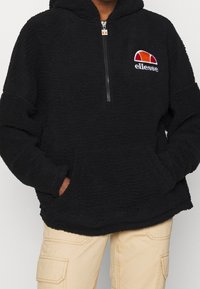 Sort fleece hættejakke med halvlynlås fortil, kengurulommer og et broderet logo på brystet. Har et blødt, tekstureret materiale.