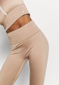 Leggings atletiche beige con vita alta. Le caratteristiche includono una texture a costine e loghi dorati sottili, che accentuano il design elegante.