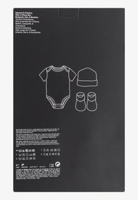 Súprava Nike s tromi časťami obsahuje biely krátky rukávový overal, bielu čiapku a biele čižmičky, všetko obrysy na čiernom pozadí. Text na obale.