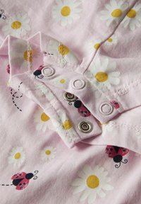 Tela de color rosa claro con un diseño de margaritas blancas y patrones de mariquitas, con botones a presión y detalle de costura visible cerca del cuello.