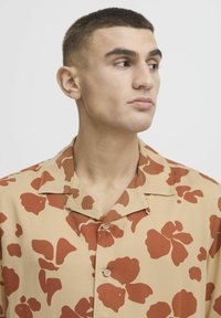 Camisa de manga corta en tejido color beige con grandes patrones florales en color óxido, con un cuello tipo camp y una abertura con botones en la parte delantera.
