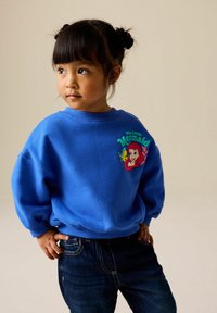 Sweat-shirt bleu avec des poignets côtelés, arborant un graphique coloré de "La Petite Sirène" sur le devant. Associé à un jean en denim foncé.