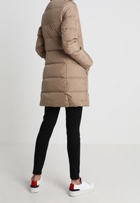 Beige prošívaný kabát s horizontálním švem, dlouhými rukávy a bočními kapsami; v kombinaci s černými kalhotami a bílými teniskami s červenými akcenty.