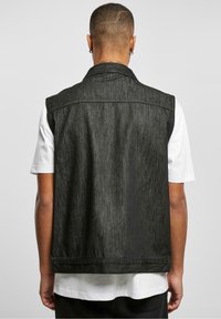 Svart denimväst med krage, öppen front och ärmlös design. Bärs över en vit T-shirt som visar en texturerad tyg.