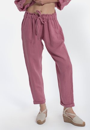 Kobieta w różowych spodniach z wysokim stanem i sznurkiem oraz beżowych sandałach espadryl na koturnie z paskami na kostkę na białym tle.