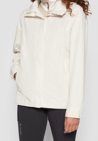 Veste légère de couleur crème avec col montant, fermeture zippée à l'avant et poches latérales. Fabriquée en matériau résistant à l'eau.