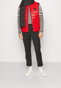 Röd pufferväst med horisontella sömmar, över en svart och vit randig långärmad tröja. Svarta byxor och vita sneakers fulländar looken.