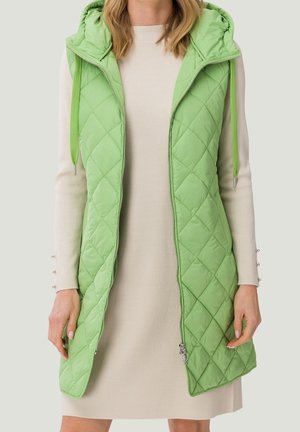 Woman wearing a light beige long-sleeve dress and a sleeveless light green quilted hooded vest with zipper open.