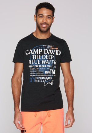 Schwarzes Baumwoll-T-Shirt mit verschiedenen weißen und blauen Textgrafiken, Rundhalsausschnitt, kurzen Ärmeln und relaxed Fit. Kombiniert mit leuchtend orangefarbenen Shorts.