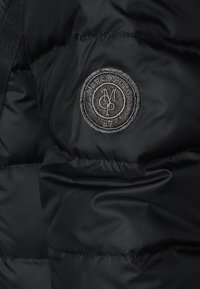 Chaqueta acolchada negra con un logo bordado redondo que dice "MARC O'POLO" y el número "67". Textura suave y lisa con un diseño acolchado.
