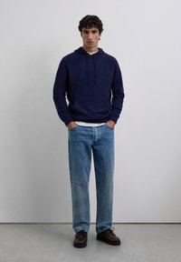 Young man standing with hands in pockets wearing navy hoodie, light blue jeans, and dark brown shoes against plain gray background.