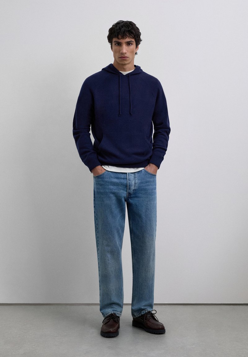 Young man standing with hands in pockets wearing navy hoodie, light blue jeans, and dark brown shoes against plain gray background.