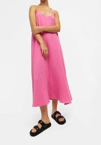 Robe longue rose sans manches en tissu texturé, avec un décolleté en V et une longueur mi-mollet, associée à des sandales plateformes noires.