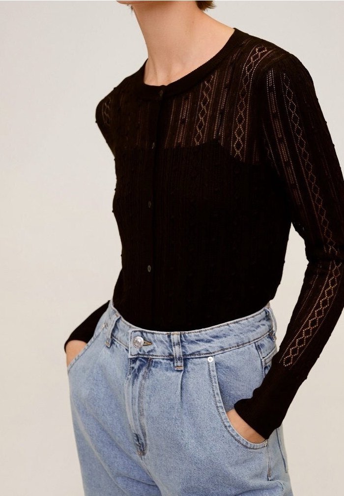 Personne portant un haut noir transparent à manches longues boutonné avec des détails à motifs, rentré dans un jean en denim bleu clair taille haute.