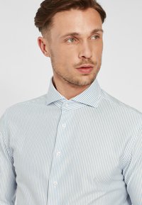 Camicia a righe blu chiaro con colletto button-down, placca dei bottoni orizzontale e tessuto liscio. Presenta bottoni bianchi e righe sottili.