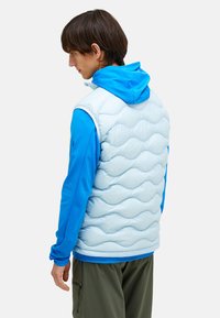 Hellblaues, puffy Weste mit Wellenmuster und weißen Akzenten, kombiniert mit einem blauen Langarmshirt, vor einem neutralen Hintergrund.