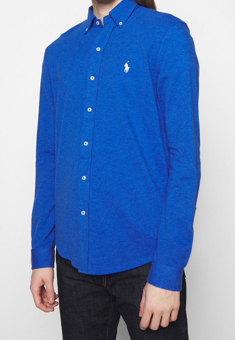 Homme portant une chemise boutonnée bleu vif à manches longues avec un petit logo blanc de cheval sur la poitrine, associée à un pantalon foncé.