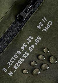 Olivengrøn stof med en glat tekstur; sort lynlås; vanddråber på overfladen; hvidt trykt tekst med numeriske oplysninger og koordinater.