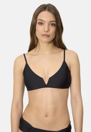 Mujer con cabello largo y castaño lleva un bikini negro con escote en V y braguitas a juego sobre un fondo claro y liso.