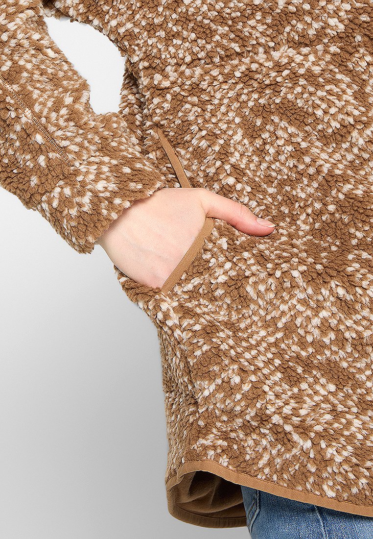 Main dans la poche d'une veste en polaire texturée marron et crème à pois, avec des bordures beiges, portée sur un jean bleu.