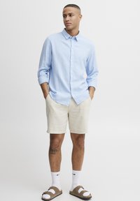 Lys blå skjorte med knapp, beige shorts, brune sandaler, hvite sokker. Modellen står med hendene i lommene og viser frem uformell sommerdrakt.