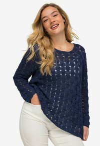 Marineblaue, weit geschnittene Strickjacke mit einem strukturierten, offenen Häkelmuster, langen Ärmeln und einem runden Ausschnitt. Kombiniert mit weißen Hosen.