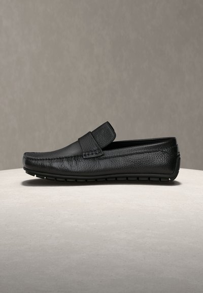 Mocassino da uomo in pelle nera con finitura testurizzata, design senza lacci e suola in gomma antiscivolo, esposto su una superficie beige con sfondo neutro.