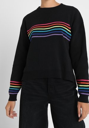 Schwarzer Pullover mit horizontalen Regenbogenstreifen über der Brust und an den Ärmelbündchen, getragen mit schwarzer Hose, gezeigt von Hals bis Hüfte.